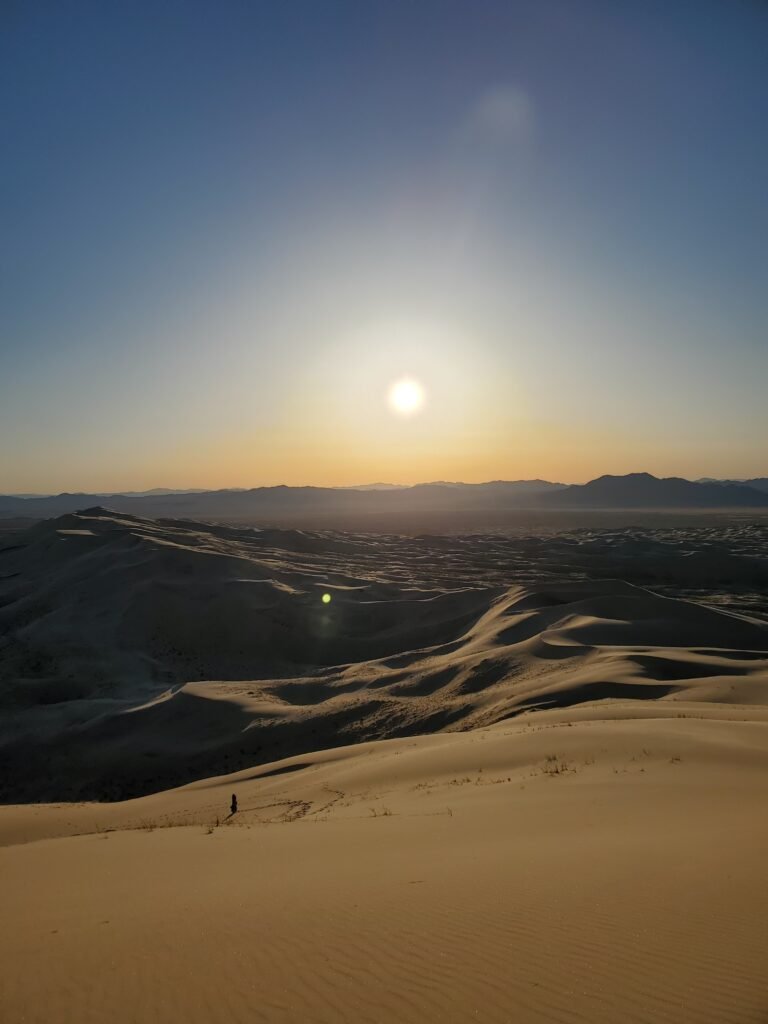a view of the desert at sunset, the sun glowing over sand dunes and shadows. in the distance, a lone figure makes footsteps in the sand